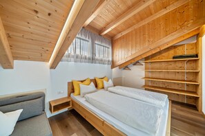 Un dormitorio con techo de madera, cama con ropa de cama blanca, sofá gris y estantería de madera.