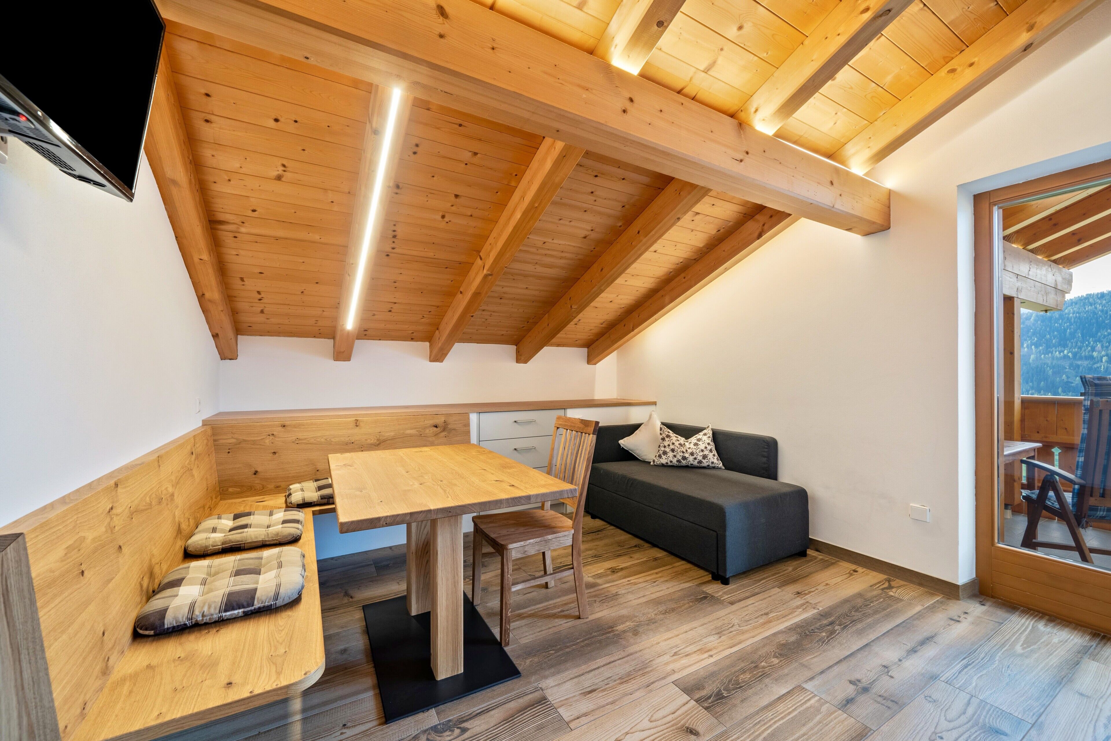 Ein Loft-Zimmer mit Holzbalkendecke, einem Esstisch mit Stühlen, einer Couch und Blick ins Freie.