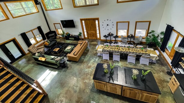 A spacious living area with a large sofa set, a dining table with chairs, and a kitchen area with cabinets. There are multiple windows providing natural light and views of the outdoors. - Black Pearl Lodge @ DeGray. Small event venue or fun family getaway! (Bismarck)