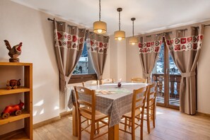 A dining area with a table set for four, wooden chairs, a wooden shelf with toys, and curtains with a patchwork design.
