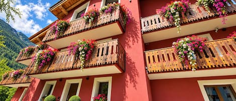 A multi-story building with balconies adorned with flowers, a red facade, and a mountainous backdrop.