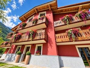 Un edificio de varios pisos con balcones decorados con flores, fachada roja y montañas al fondo.