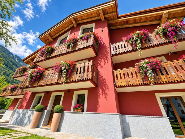 A multi-story building with balconies adorned with flowers, a red facade, and a mountainous backdrop.
