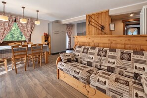 Una sala de estar acogedora con un sofá estampado, una mesa de comedor de madera, sillas y una zona de cocina con armarios de madera y un tendedero.