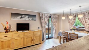 Un salon comprenant un téléviseur, un meuble en bois, une table à manger, des chaises et une grande fenêtre avec des rideaux.