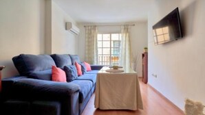 Un salón con sofá, una mesita, un televisor de pantalla plana montado en la pared y una ventana con cortinas.