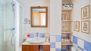 Un baño con ducha, lavamanos, espejo y estantes con fotografías enmarcadas.
