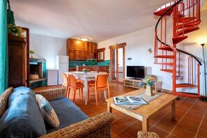 A living room with a sofa, a coffee table, and a TV. There is a kitchen area with wooden cabinets and a red spiral staircase.