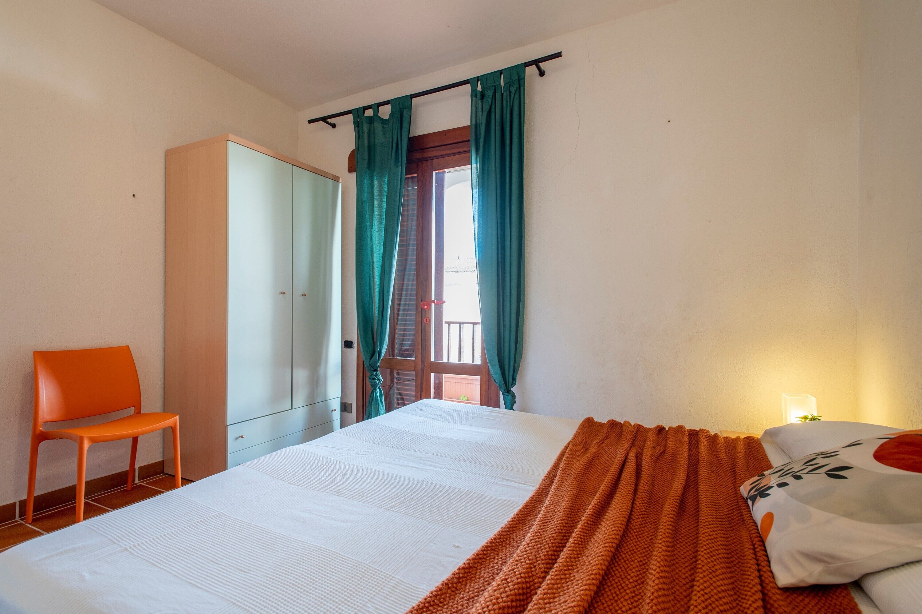 Una habitación con una cama, una silla naranja, un armario y una ventana con cortinas.