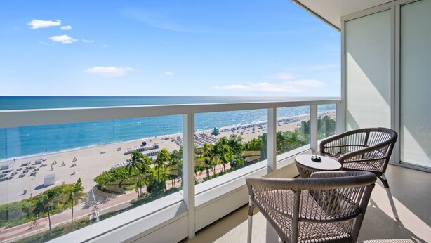 A balcony with wicker chairs and a small table, overlooking a beach with umbrellas and palm trees. - Direct Ocean View Studio at Sorrento Fontainebleau, Miami Beach (Miami Beach)