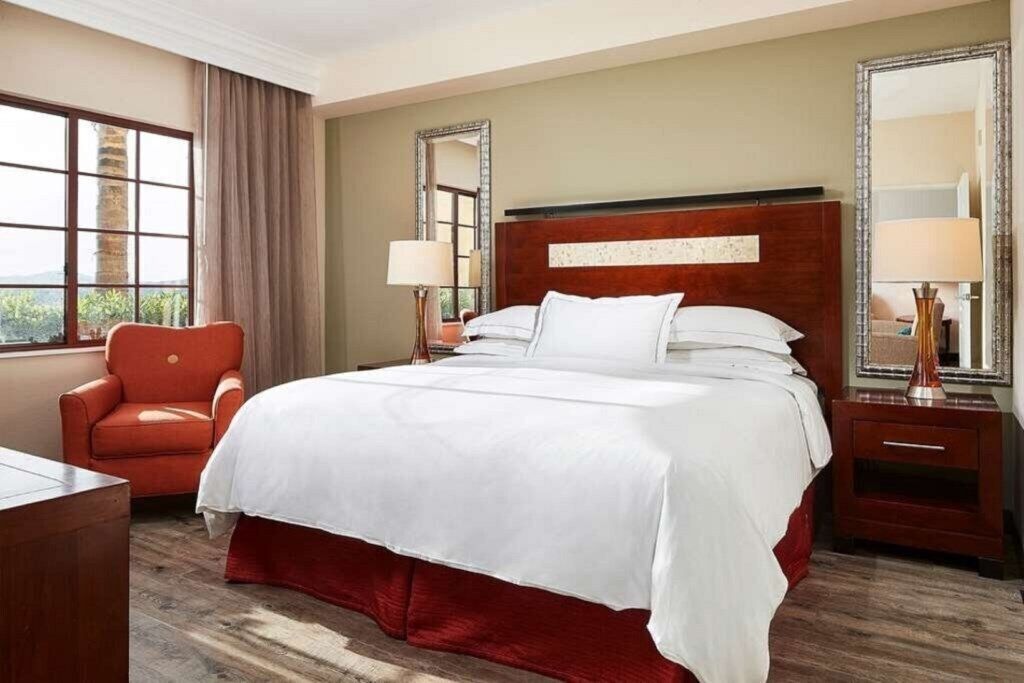 A hotel room with a large bed, a red chair, two bedside tables with lamps, and a large window with a view.