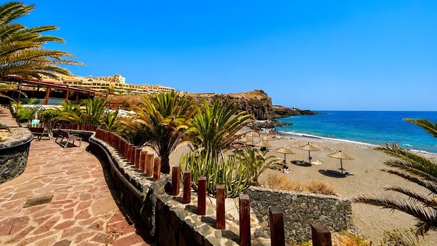 Un complejo turístico costero con un paseo pavimentado, palmeras, sombrillas de playa y vistas al mar y edificios al fondo.