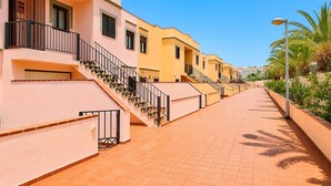 Una calle residencial con aceras de tejas de barro, balcones con barandillas negras y una hilera de casas con fachadas rosa pálido y amarillo.