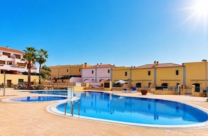 A swimming pool area with a clear sky, palm trees, and buildings with balconies.