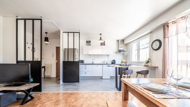 Een moderne keuken met witte kasten, een eethoek met een houten tafel, en een groot raam met gordijnen.