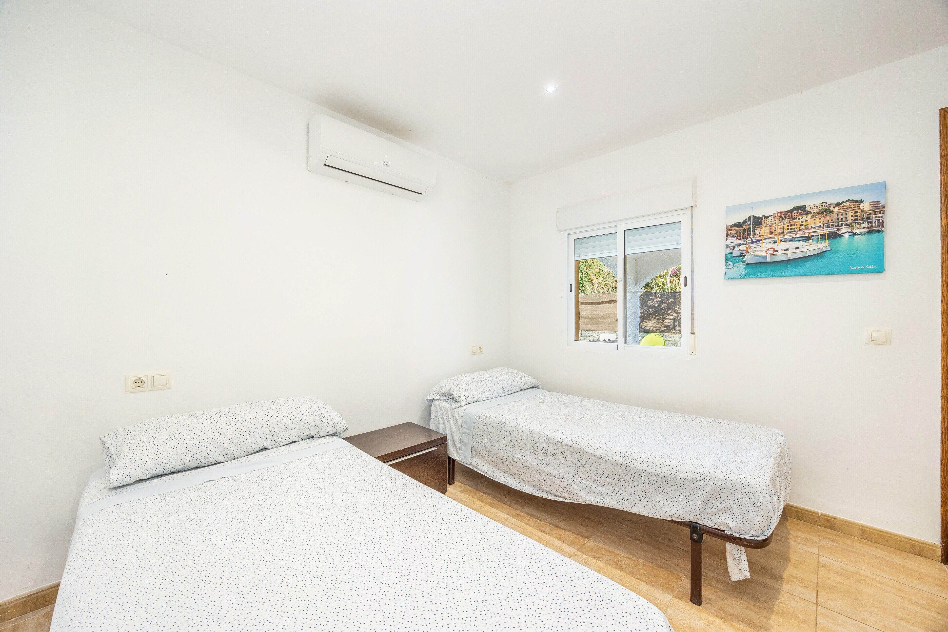 Un dormitorio con dos camas individuales, un equipo de aire acondicionado, un ventanal con vistas a un pueblo costero y un cuadro en la pared.