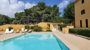 A swimming pool with lounge chairs, a yellow building, and a forested hill in the background. - YourHouse Sol i Mar 1 (Cala Mesquida)