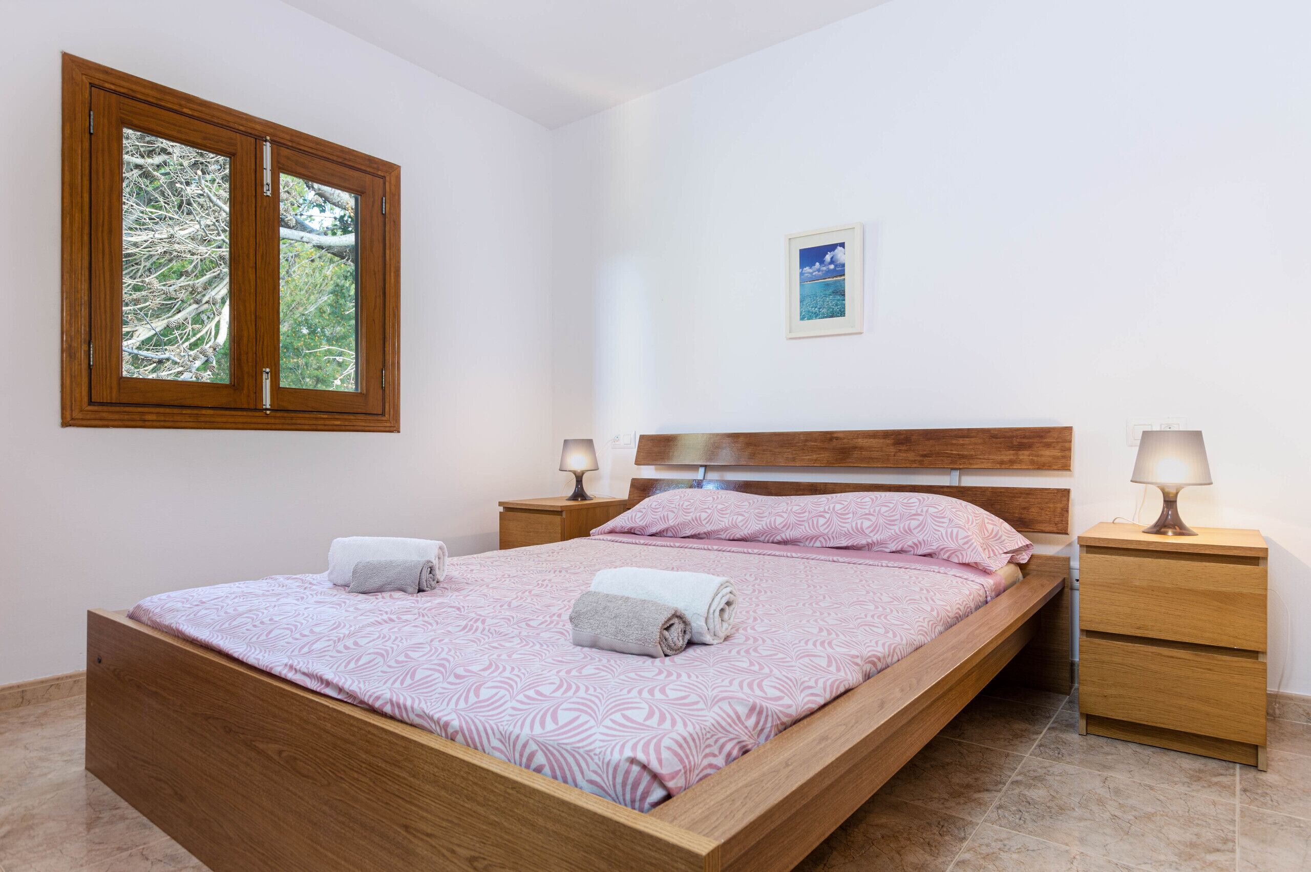 A neatly made bed with a patterned bedspread, two bedside lamps, a small bedside table, and a window with a view of trees.