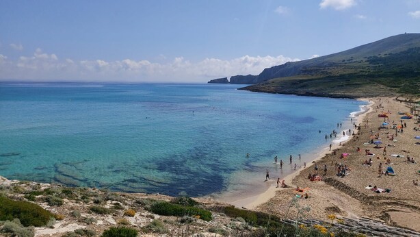 Una platja amb aigua blava i cristal·lina, sorra daurada i un paisatge amb zones muntanyoses al fons.