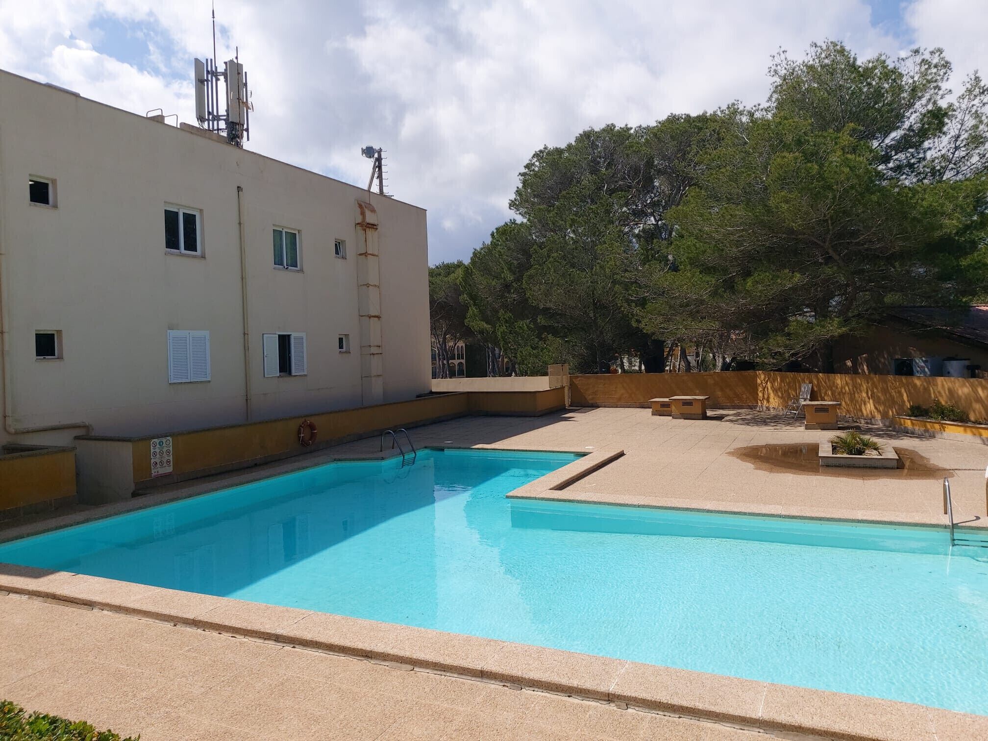A swimming pool with a clear blue water, surrounded by a beige building with windows and a lifebuoy.