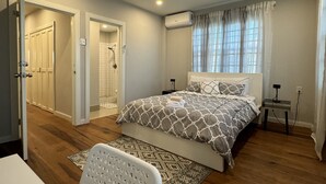 A bedroom with a bed, a desk, and a chair. There is a bathroom with a shower and a toilet. The room has wooden flooring and white walls. - A modern Townhome at Farm. 5 minutes drive from the Providence National Stadium (East Bank Demerara)
