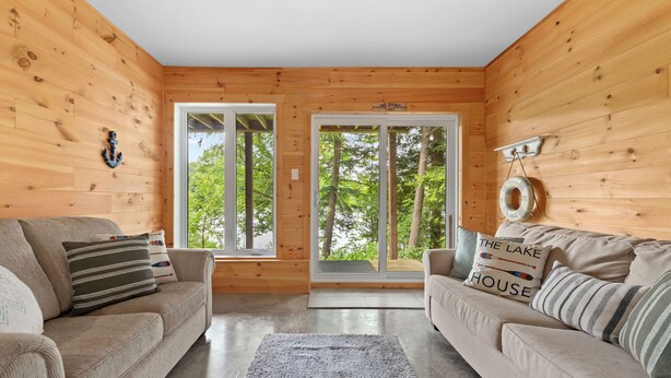 Ein gemütliches Blockhaus-Interieur mit zwei Sofas, einem Teppich, Holzwänden und großen Fenstern mit Blick auf Bäume.