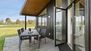 Un espace extérieur abrité, aménagé pour les repas, avec une table et des chaises, de grandes portes vitrées et une vue sur un champ verdoyant.