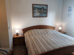 A neatly arranged hotel room with a bed, bedside lamps, a nightstand, a small table, and a framed painting on the wall.