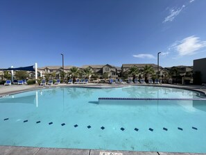 Una piscina con marcas azules para carriles, rodeada de sillas de descanso y palmeras, con edificios residenciales al fondo.