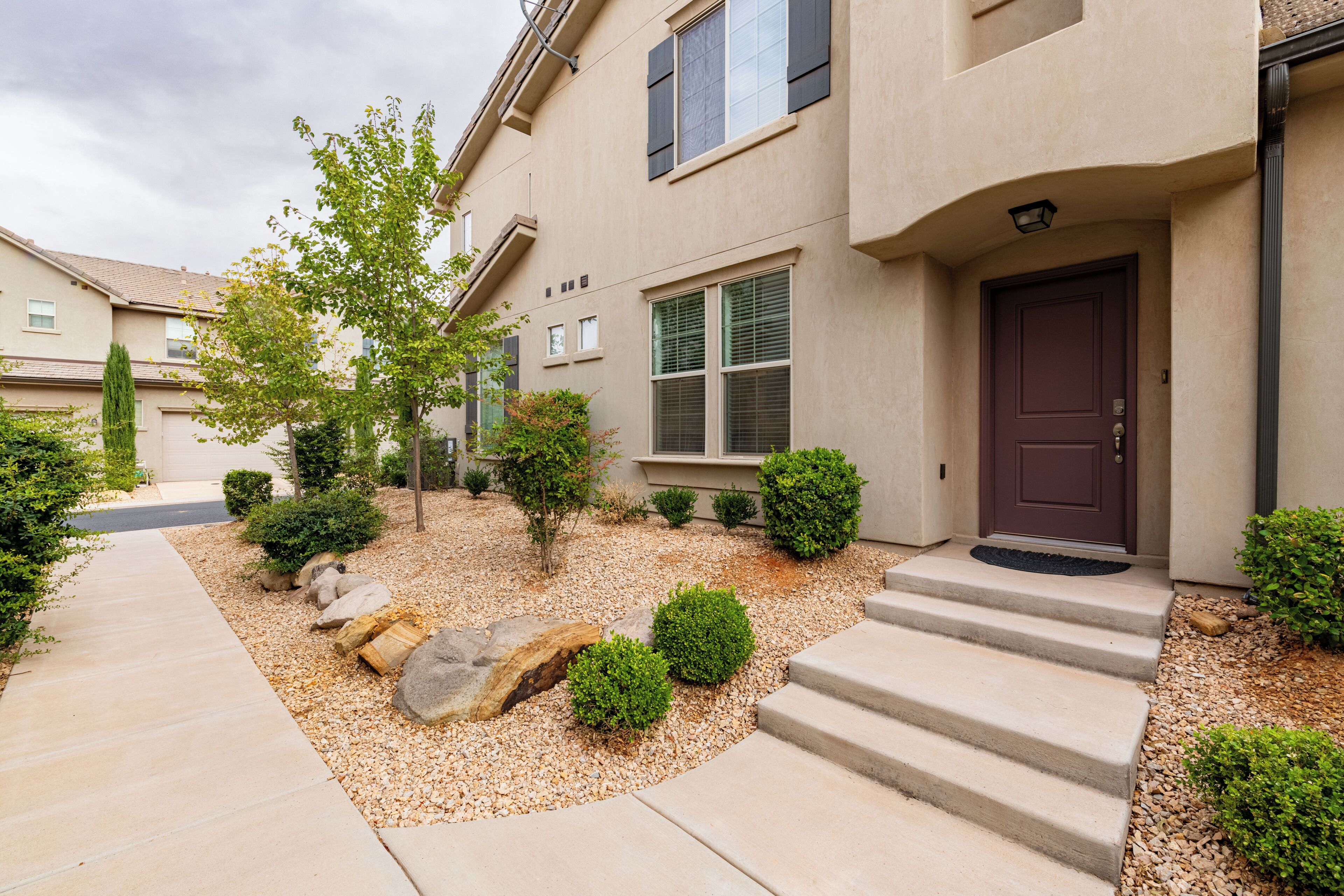 A modern two-story house with a beige exterior, a dark brown front door, and a small front yard with a gravel pathway, steps, and various shrubs.