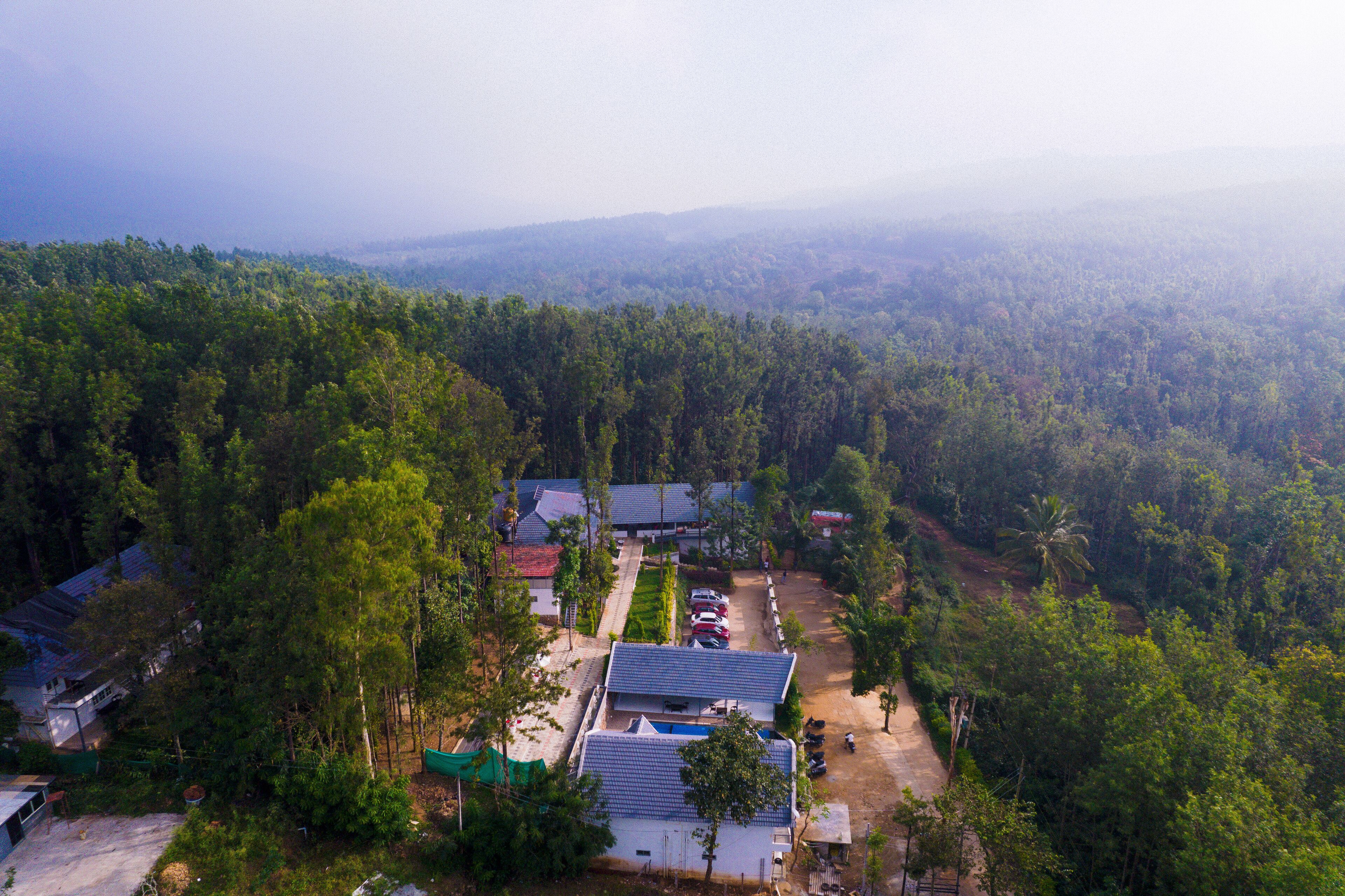 An aerial view of a residential area with multiple houses surrounded by dense forest. There are visible pathways and a few parked cars.
