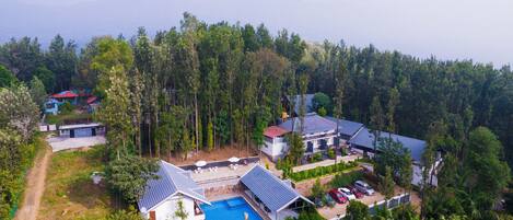 An aerial view of a resort with multiple buildings, a swimming pool, and a forested backdrop.