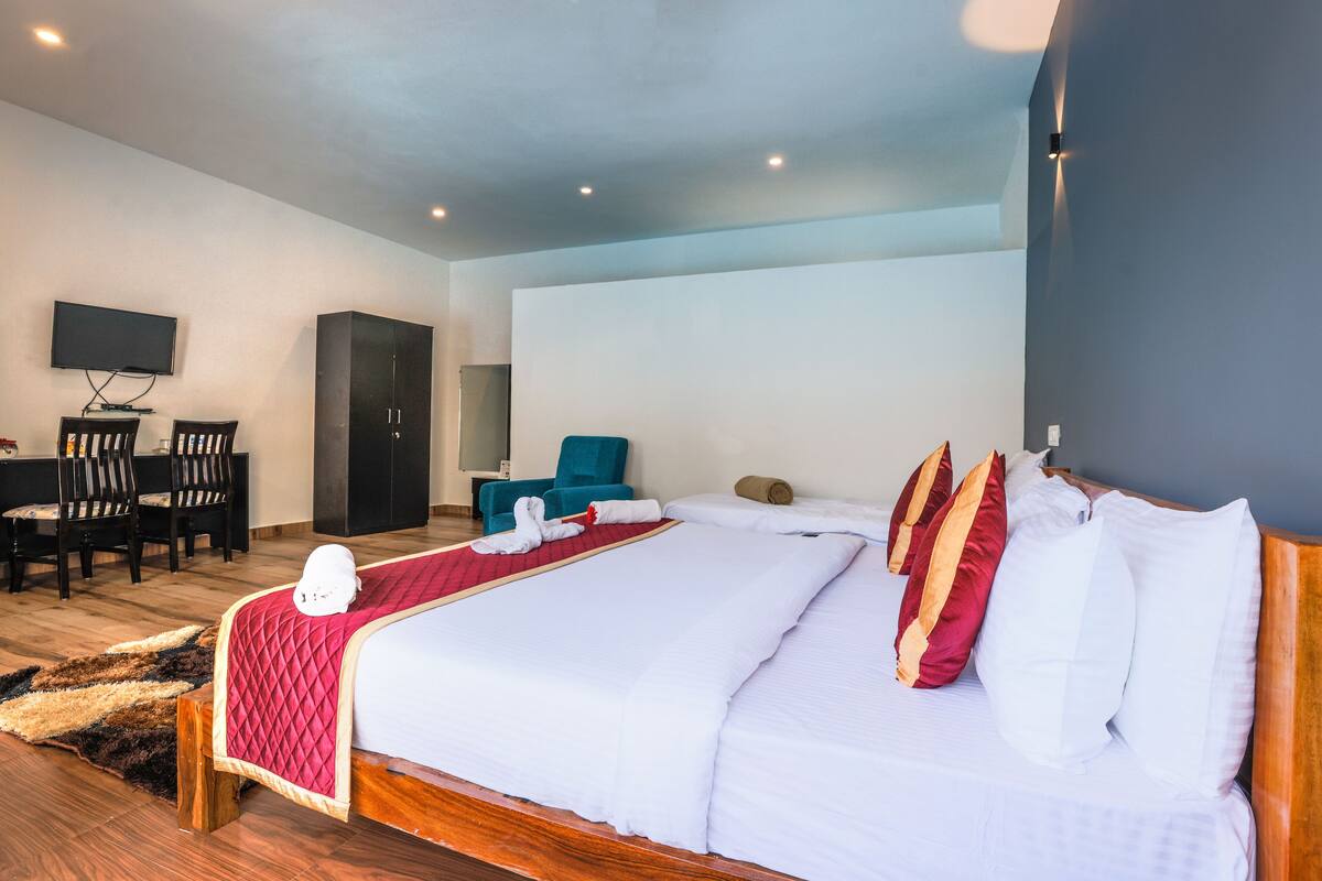 Quarto de hotel com uma cama grande, uma escrivaninha com cadeiras, uma televisão e uma poltrona azul.