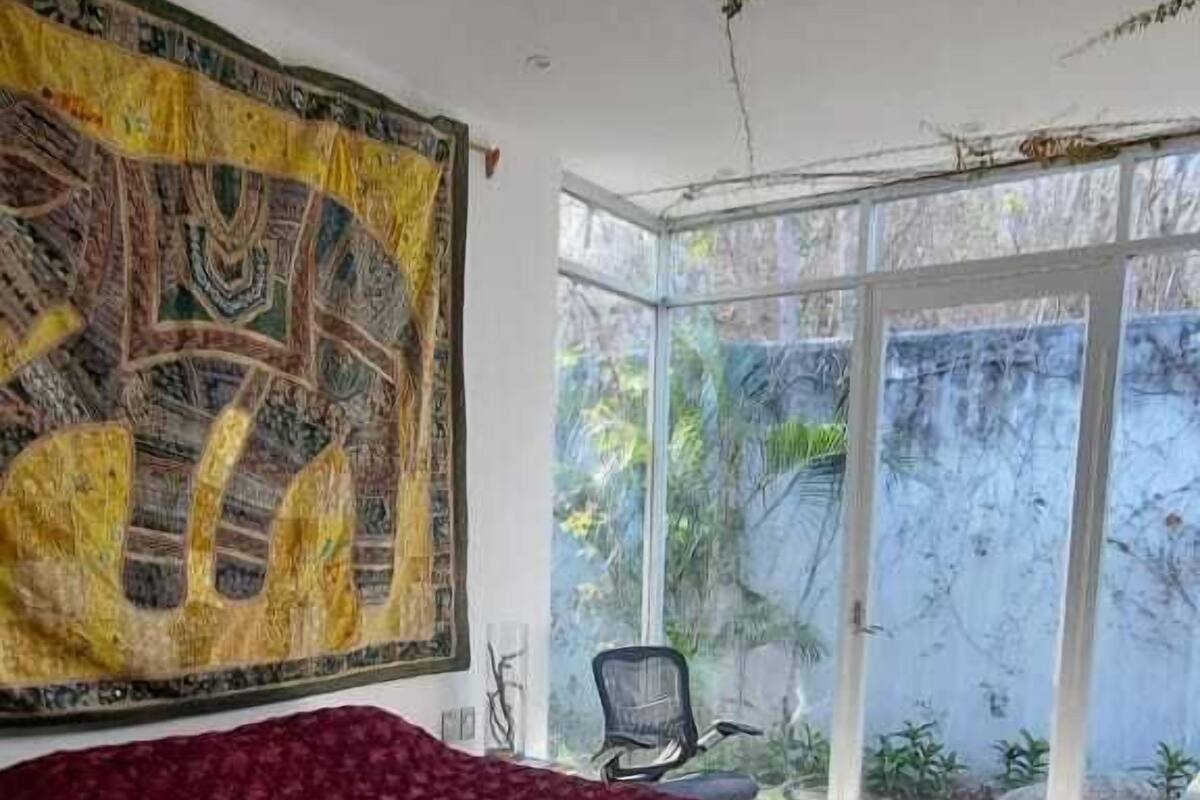 A bedroom with a large bed, a red bedspread, a patterned tapestry, and a view of greenery through a glass door.