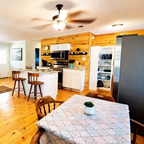 A kitchen with wooden walls, a central island, bar stools, a dining table with a patterned tablecloth, and a refrigerator. - Knee Knocker at Caddo Lake - Hot tub - Firepit  - Nature Dock (Karnack)