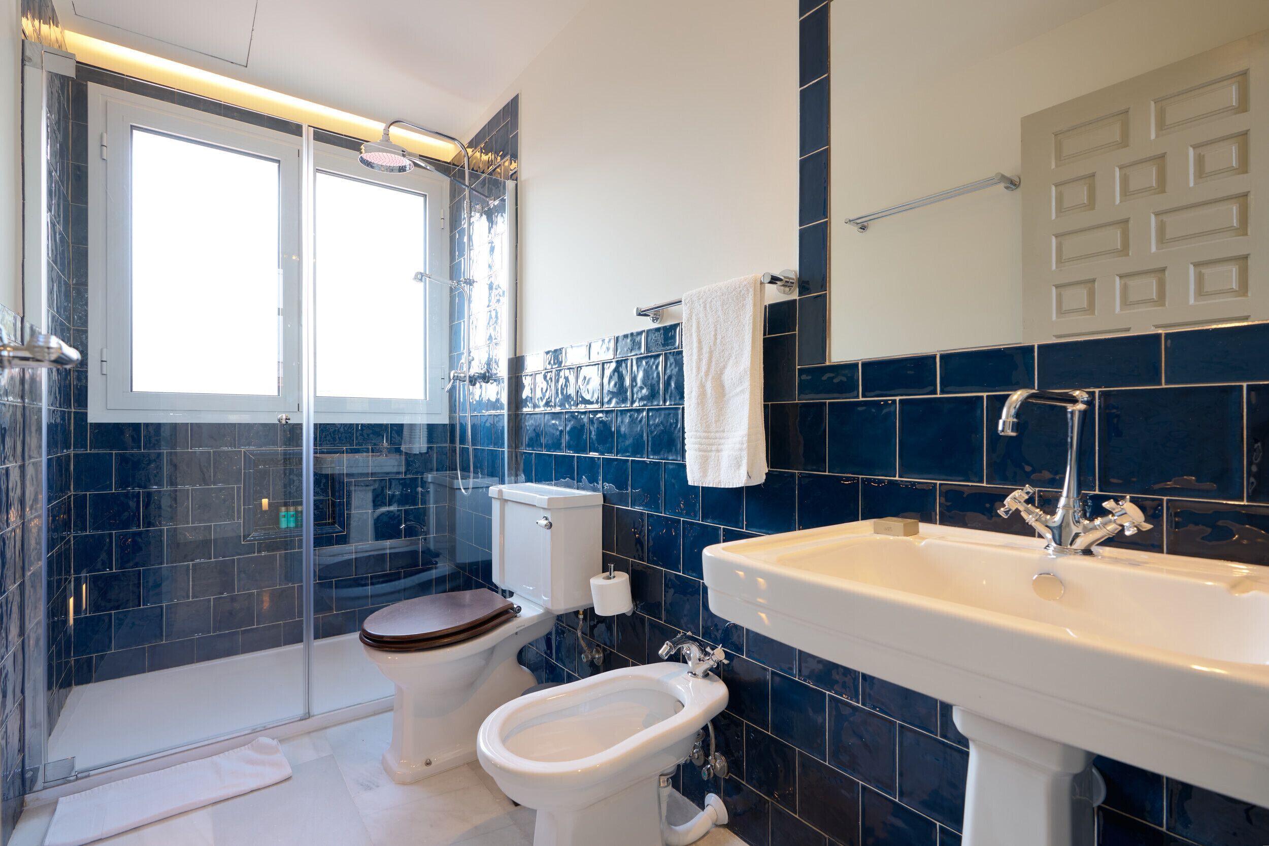 A modern bathroom with a glass-enclosed shower, a toilet with a wooden seat, a bidet, and a large sink with a mirror above it.