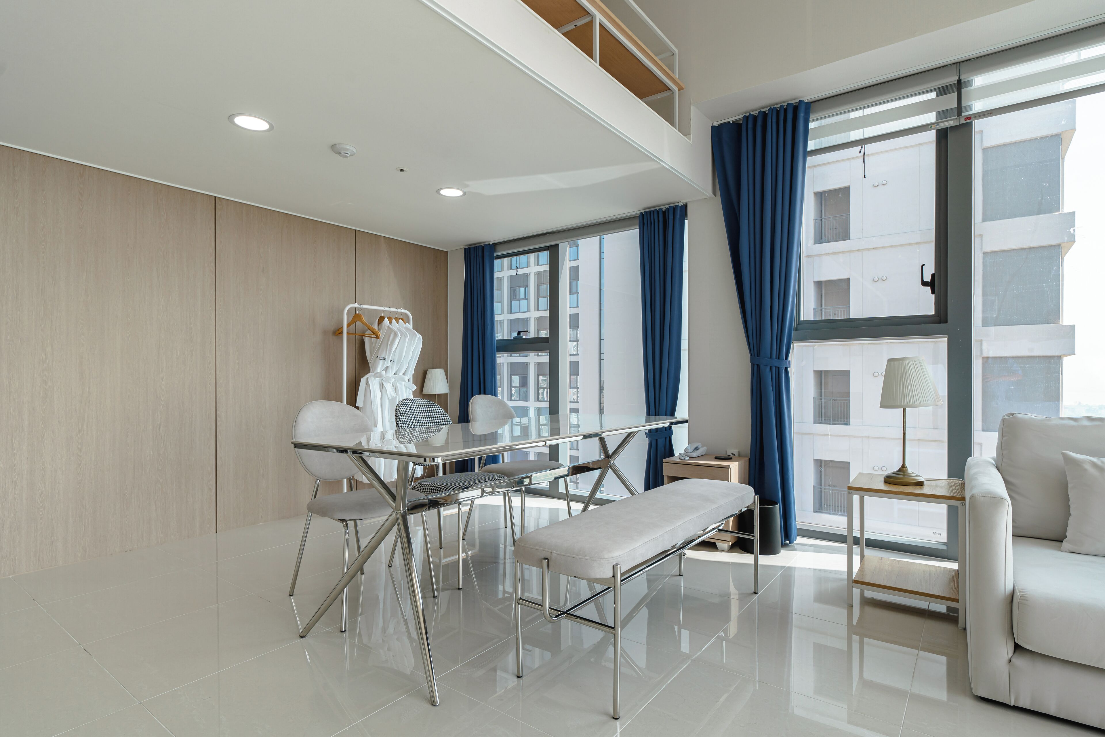 A modern hotel room with a dining area, a sofa, a bench, and a large window with curtains.