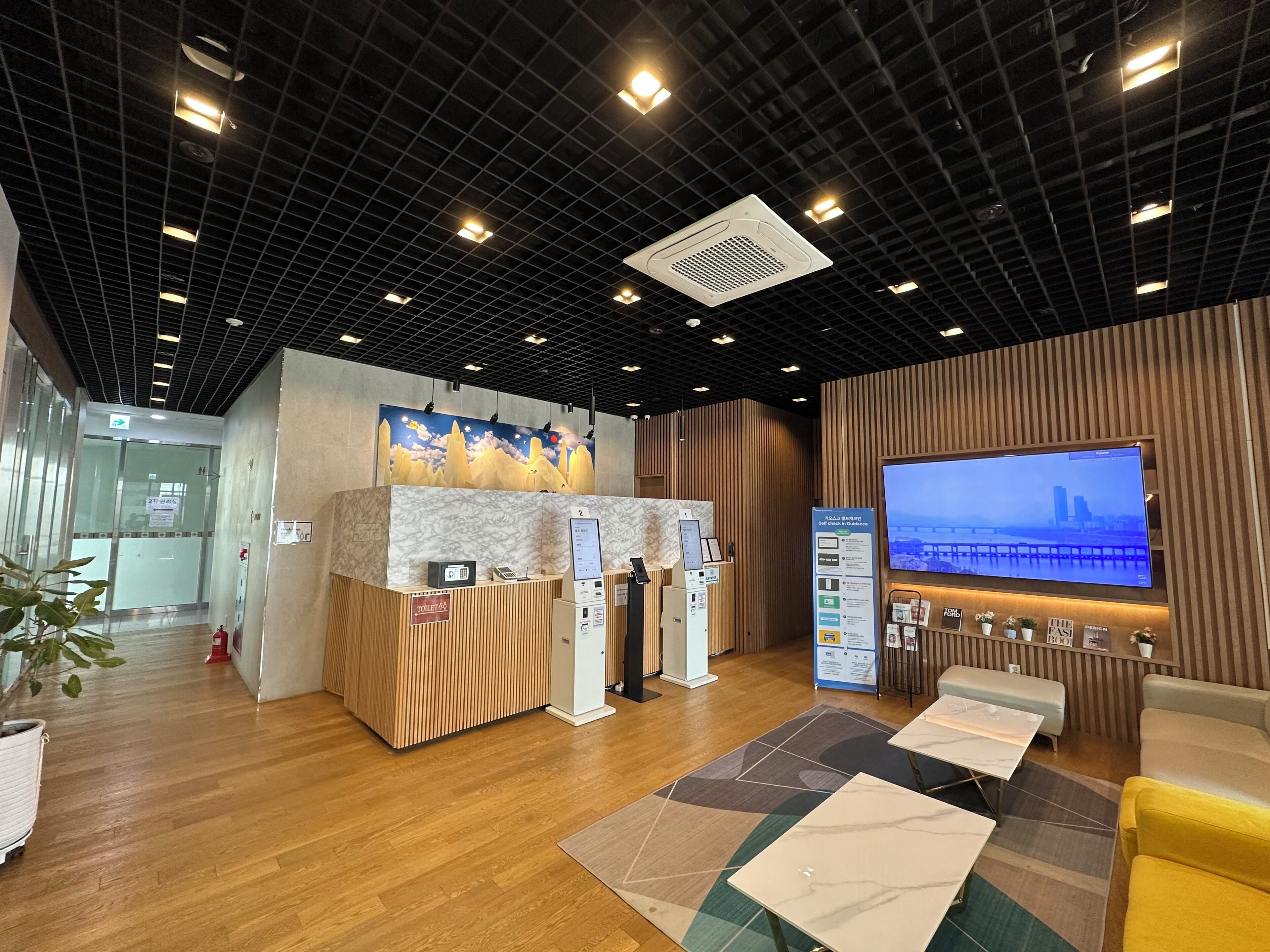 Zona de recepció amb mostrador de marbre, panell informatiu digital, una gran pantalla de televisió i taulell de fusta.