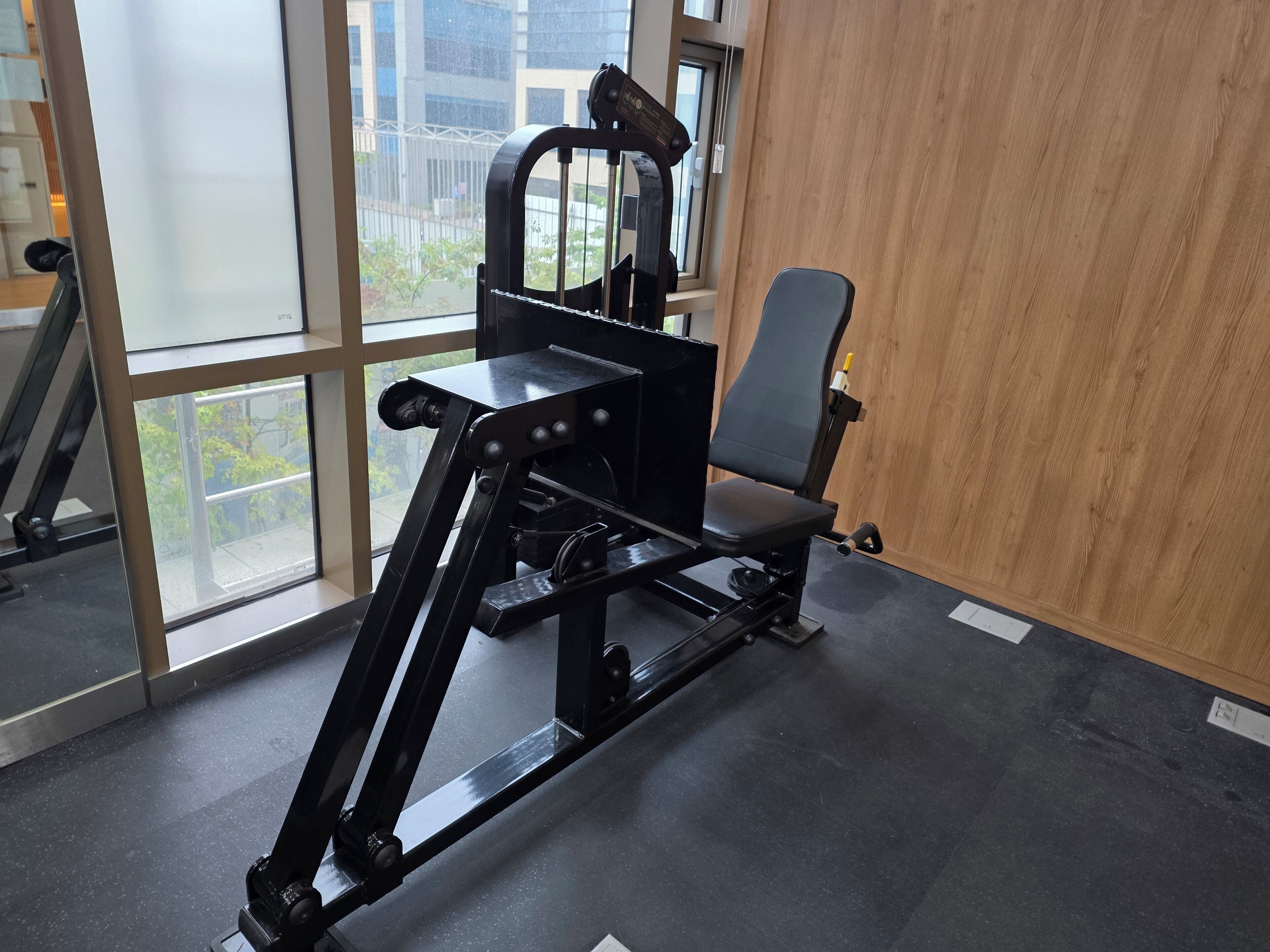 Ein Fitnessraum mit einer schwarzen Trainingsmaschine, großen Fenstern und einer Holzwand.