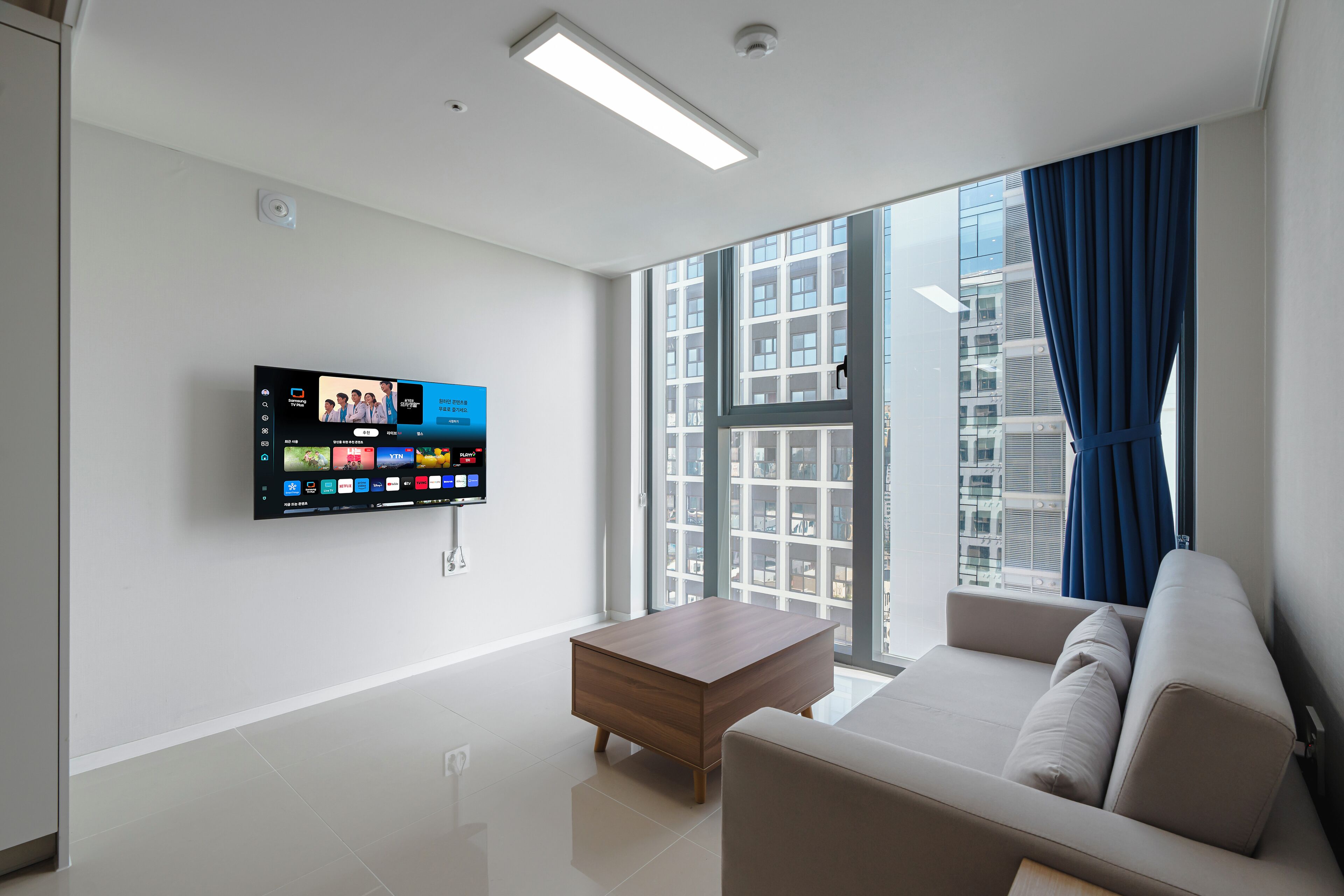 A modern living room with a flat-screen TV, a sofa, a coffee table, and a large window with curtains.
