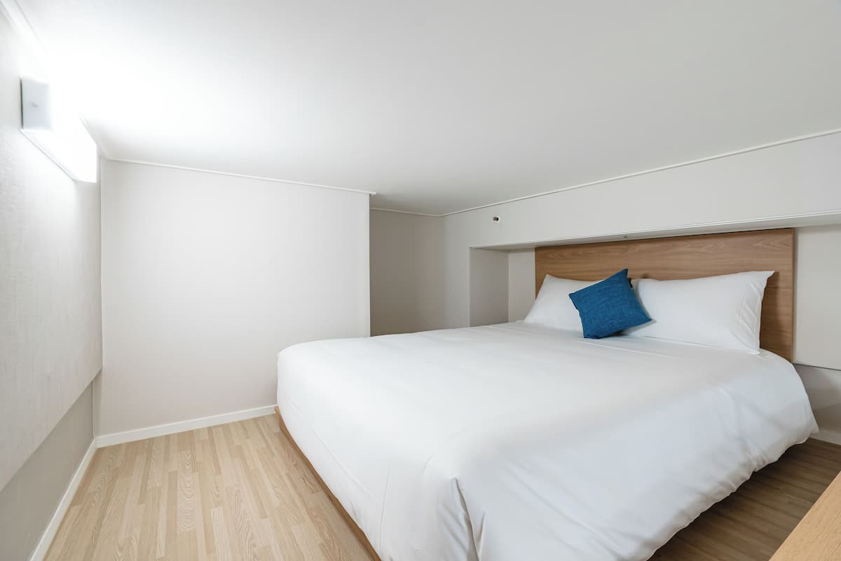 A neatly made bed with white linens and a single blue pillow, a wooden headboard, and a light-colored wooden floor.