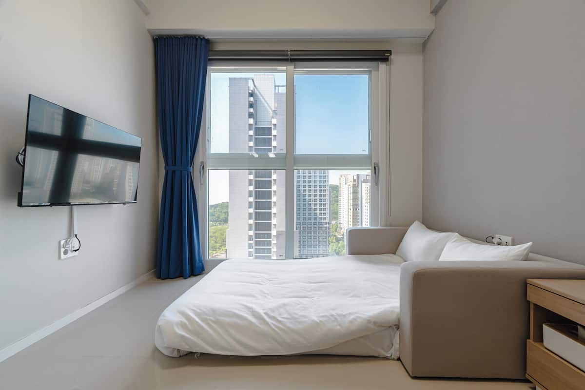A modern hotel room with a large bed, a TV mounted on the wall, and a view of tall buildings through the window.