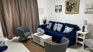 Una sala de estar con un sofá azul, dos sillones grises, una mesa de centro blanca y una pequeña mesita auxiliar con una lámpara.