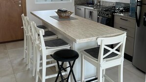 Cocina compacta con una isla central, taburetes blancos, estufa y refrigerador.