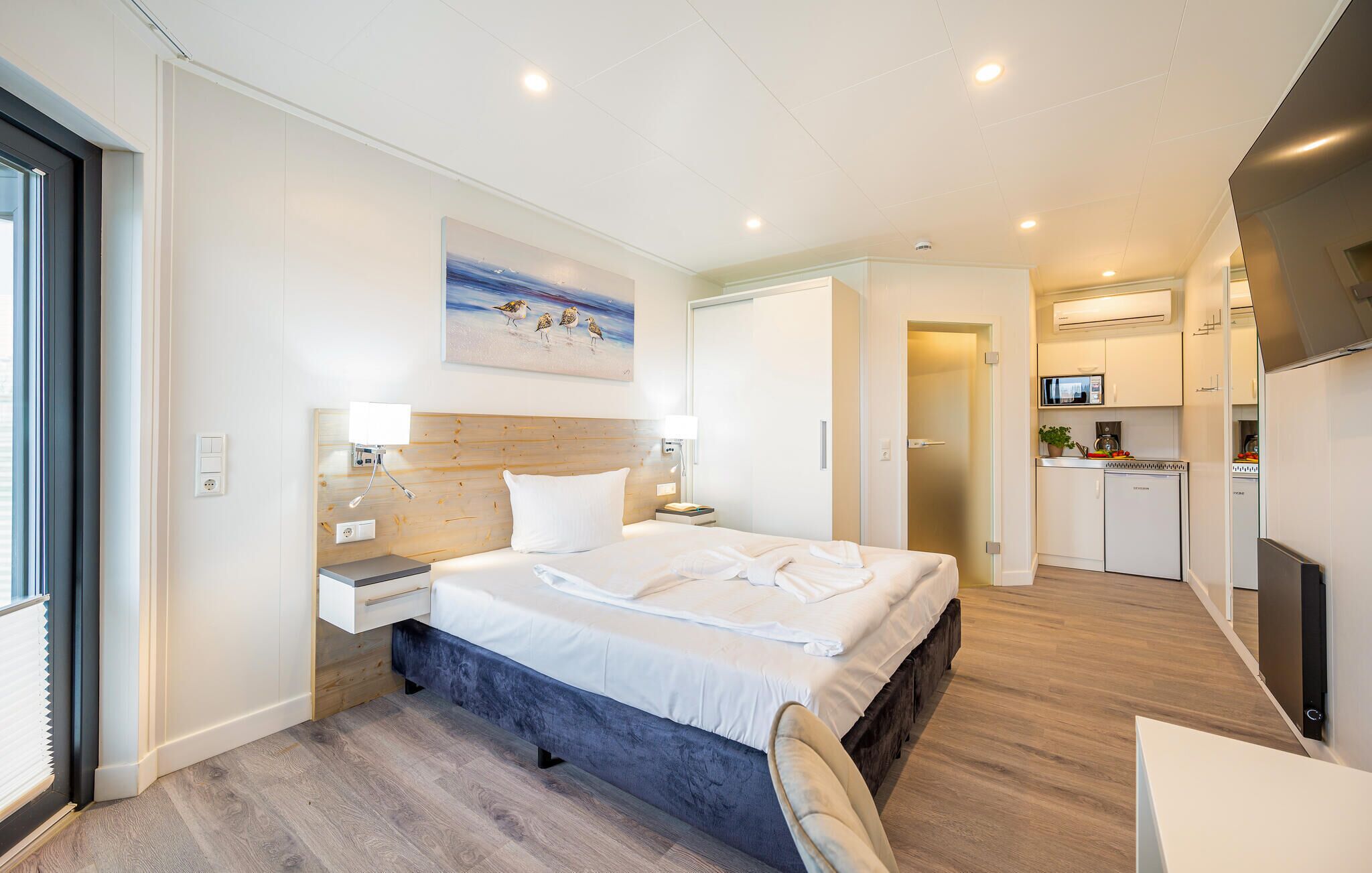 A modern hotel room with a large bed, bedside tables, a desk, and a kitchenette.