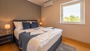 A neatly made bed with a duvet, pillows, and a blanket. There is a bedside table with a lamp, a window with a view of greenery, and an air conditioning unit on the wall.