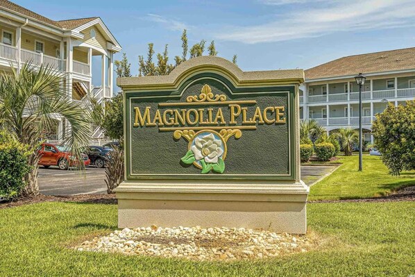 A sign for Magnolia Place with a decorative emblem, surrounded by well-maintained grass and a parking area.