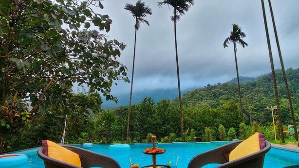 A poolside setting with wicker chairs, a glass table with food and drinks, and a view of a lush green landscape with palm trees. - Hill Crest Resort (Madikeri)