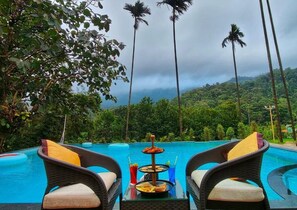 A poolside setting with wicker chairs, a glass table with food and drinks, and a view of a lush green landscape with palm trees. - Hill Crest Resort (Madikeri)