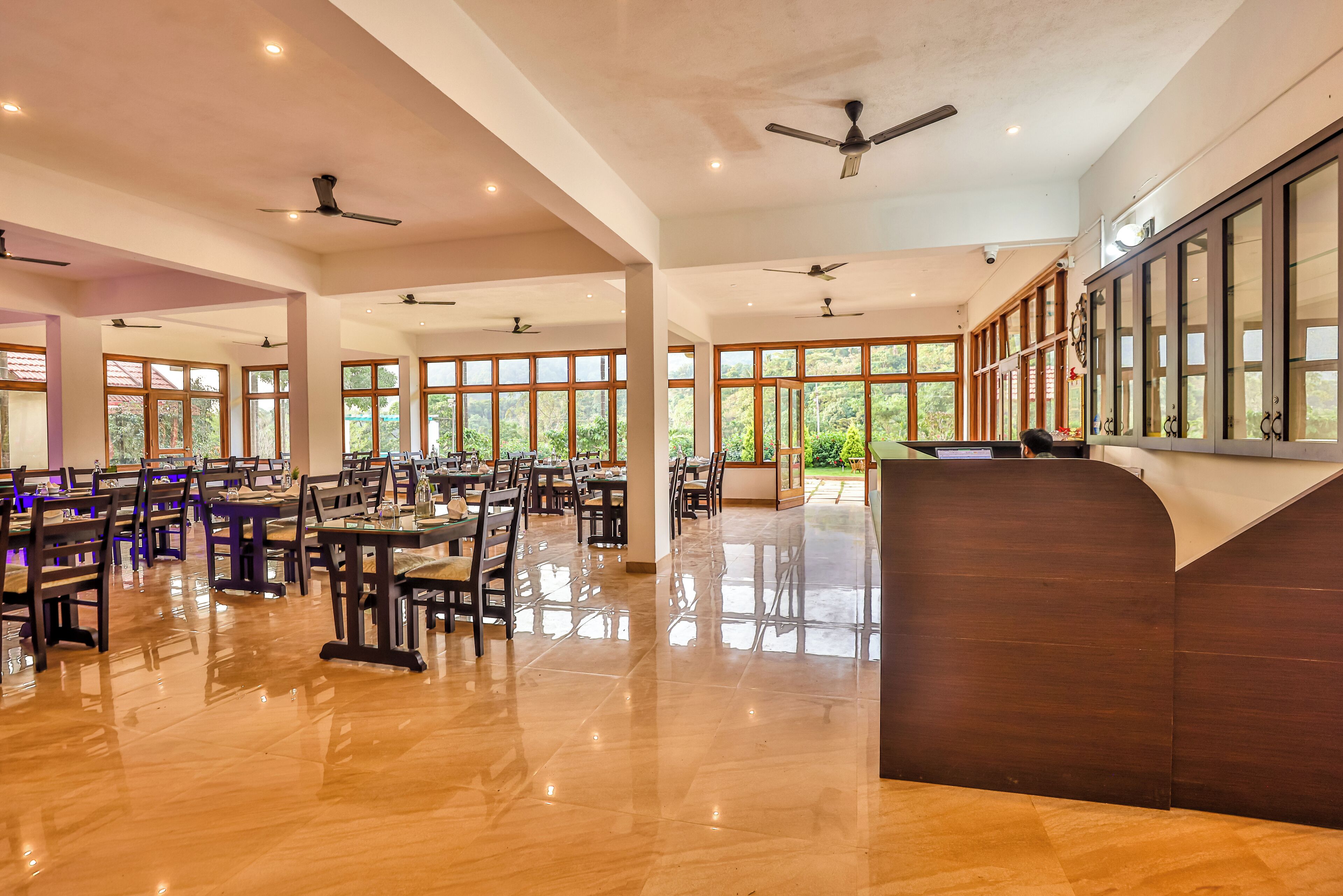 Un vaste espace dédié à la salle à manger, avec de nombreuses tables et chaises, des ventilateurs de plafond et de grandes fenêtres offrant une vue sur la verdure.